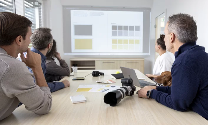 So arbeiten wir: von der Idee zum Ergebnis Team im Meeting an einem Besprechungstisch mit Kamera und Laptop, auf der Leinwand Entwürfe für ein Farbkonzept